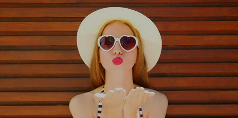 Portrait of young woman blowing her red lips sending sweet air kiss wearing a summer straw hat on a background