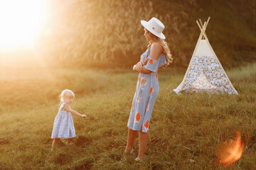 Beautiful mother with her little daughter having fun near wigwam in the field. Spending time together, outside, on vacation, outdoors. Beautiful sunset light in the garden or in the park. © Andriy Medvediuk
