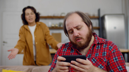 Aged mother scolding adult son sitting at home on sofa using mobile phone