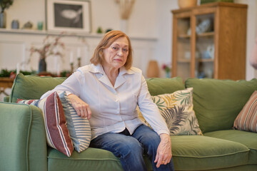 Portrait of an elderly woman sitting on a couch at old age home. Caucasian woman sitting on a sofa and looking at camera. Care for lonely pensioners.