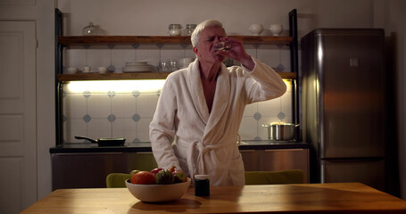 Senior man in bathrobe taking pills and drinking water in kitchen in evening