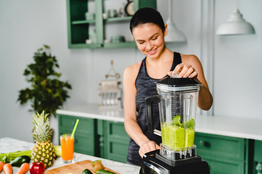 Cheerful young woman making smoothie using blender in the kitchen