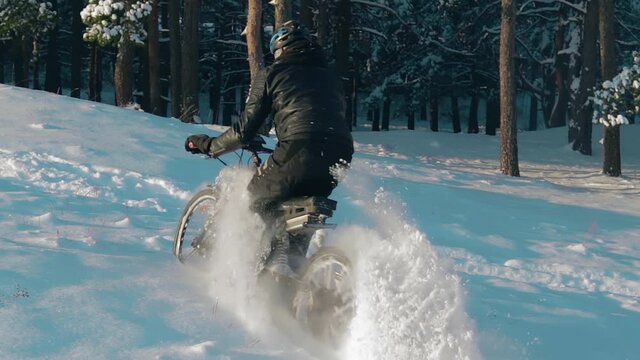 Cyclist in Black Clothes, Helmet and Snow Glasses Riding on Electric Bike in the Snow in Winter Forest. E-Bike Riding and Drifting at Cold Sunny Day - Slow Motion