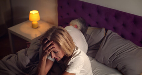 Senior woman sitting in bed and feeling stressed with husband sleeping on background