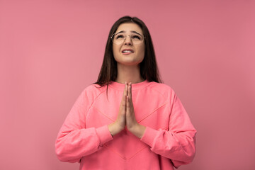 Photo of attractive young woman in glasses uncertainly praying , wears casual pink t-shirt isolated pink color background
