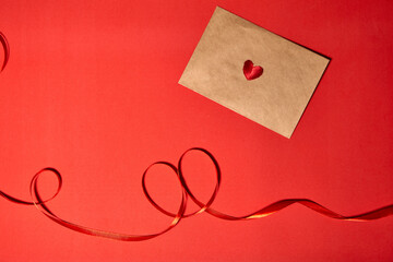 A red heart lies on a brown envelope for Valentine's Day. Red backdrop with envelope and red heart. Love letter or message concept. Flat-lay, top view. Copy space for your text.