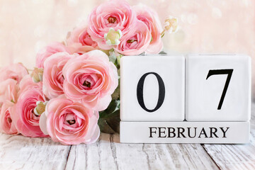 Fototapeta premium White wood calendar blocks with the date February 7th and pink ranunculus flowers over a wooden table. Selective focus with blurred background. 