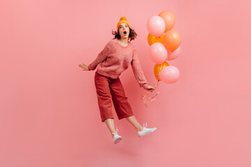 Excited young lady in hat jumping on pink background. Adorable girl with air balloons fooling...