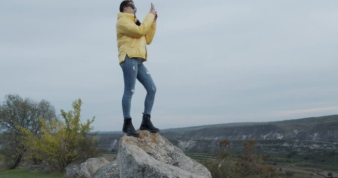 a girl with the phone in her hand takes pictures, perched on a rock in nature.