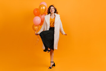 Pleased woman in coat dancing with air balloons. Attractive birthday girl having fun on yellow background.