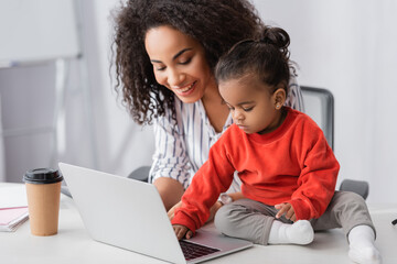 toddler african american child typing on laptop near cheerful mother