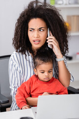 african american mother talking on smartphone while sitting with toddler daughter near laptop on blurred foreground