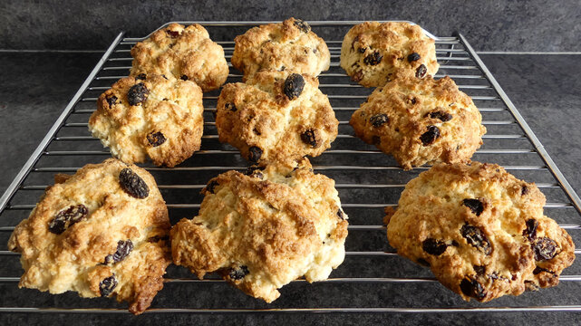 Close up of a batch of home made, freshly baked rock cakes cooling on a chrome grill standing on a black marbled surface. Interior landscape image with selective focus on middle bun. 