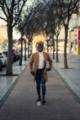 Chico negro atractivo con abrigo largo beige posando y usando el smartphone