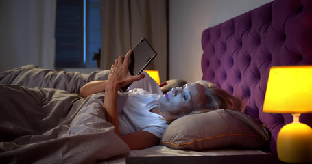 Sleepless senior woman in bed at night using mobile phone while husband sleeping