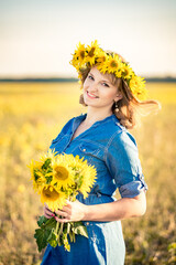 Cute redhaired smiling girl dressed in a dress-shirt made of denim in the field full of sunflowers, wreath and bouquet of sunflowers.