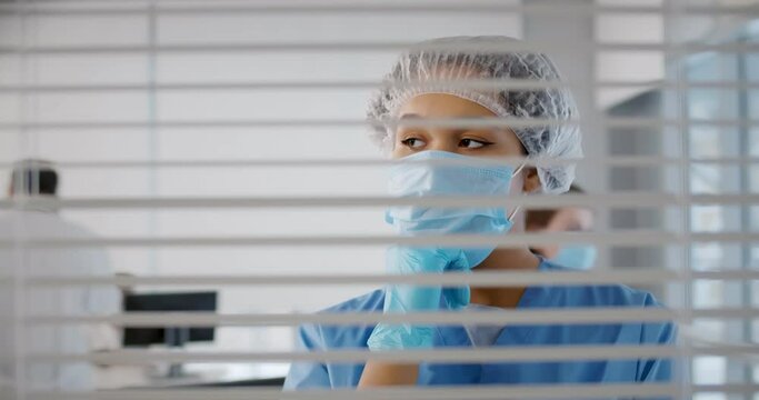 Female wearing surgeon uniform and medical mask thinking standing in office behind window with blinds