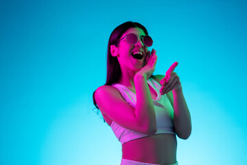 Laughting, pointing. Brunette woman's portrait on blue studio background in mixed neon. Beautiful model in stylish eyeglasses. Concept of human emotions, facial expression, sales, ad, fashion.
