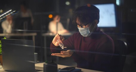 Asian businessman working in office at night wearing face mask, sitting at desk using hand sanitizer. - Powered by Adobe