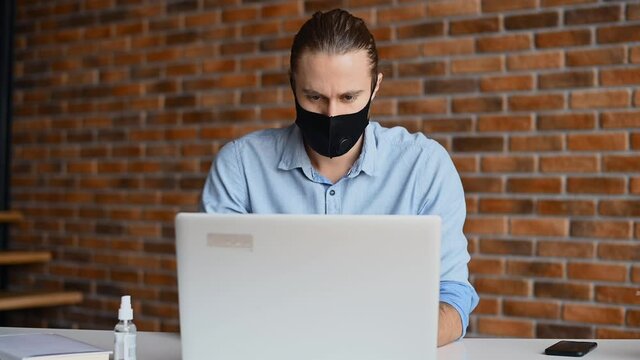 A thoughtful hipster guy wearing medical mask is using laptop for work at modern co-working. Protective measures for office staff during pandemic and virus outspread