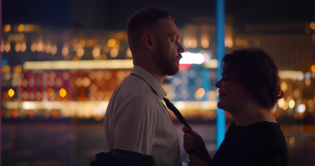 Side view of elegant woman and man flirting standing near panoramic window with night city view