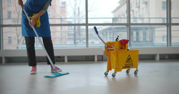 Uniformed cleaner wiping floor using mop and cart in business center
