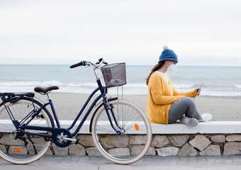 A digital marketer is working on a new laptop promotional project while sitting on the seaside promenade with bicycle. The independent woman communicates with clients.