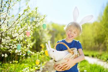 Cute boy in bunny ears holding wicker basket with white toy rabbit during hunt for colorful eggs in...