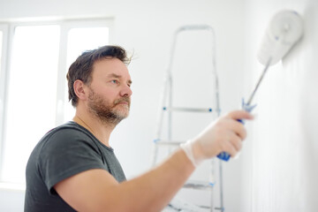 Portrait of a handsome mature man making repairs in the apartment. The person paints the wall white with roller. Do it yourself. DIY