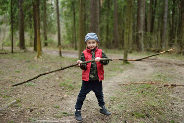 Little boy in red vest is playing with big branch and having fun in forest on early spring day. Outdoor activity for children.