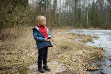 Little boy playing on shore of forest lake on early spring day. Outdoor activity for kids.