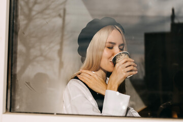 Young stylish woman at a cafe, drinking coffee, sitting by the window in sun light.