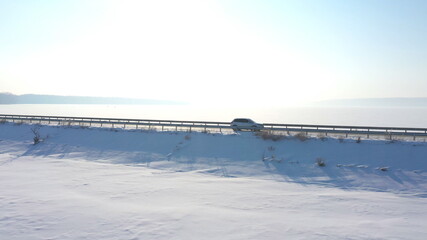 Aerial shot of white car riding through snow covered road near a frozen lake. SUV driving at countryside route near icy pond at sunny winter day. Flying over the auto moving through scenic landscape
