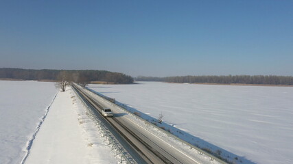 SUV driving at countryside route near icy pond at sunny winter day. Aerial shot of white car riding through snow covered road near a frozen lake. Flying over the auto moving through scenic landscape