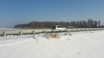 Aerial shot of car riding through snow covered road near frozen lake. White SUV driving at dam route on winter day. Flying over the auto moving through bridge of river. Scenic landscape way. Top view