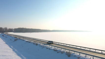 Aerial shot of car riding through snow covered road near frozen lake. White SUV driving at dam route on winter day. Flying over the auto moving through bridge of river. Scenic landscape way. Top view