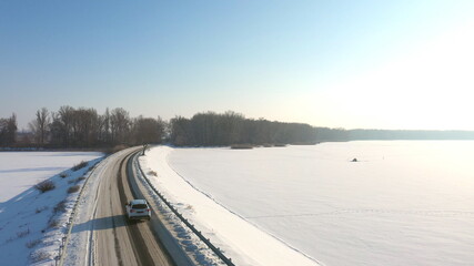 Obraz premium Aerial shot of car riding through snow covered road near frozen lake. White SUV driving at dam route on winter day. Flying over the auto moving through bridge of river. Scenic landscape way. Top view