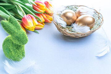 Golden easter colour eggs in basket with spring tulips, white feathers on pastel blue background in Happy Easter decoration. Spring holiday concept.