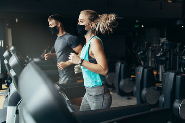 Fototapeta premium Young fit woman and man running on treadmill in modern fitness gym. They keeping distance and wearing protective face masks. Coronavirus world pandemic and sport theme.