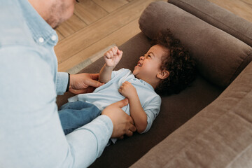 Little boy lying on sofa and laughing while father tickling him