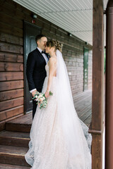 Naklejka premium Full length portrait of handsome man in stylish suit embracing his charming young bride in white wedding dress. Happy newlywed couple posing near wood house.