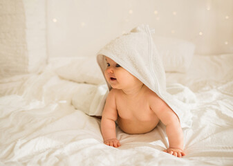 Obraz na płótnie Canvas little baby girl in a white towel with a hood is on all fours on a white blanket on the bed