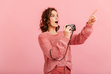 Shocked girl in sweater holding camera. Studio shot of woman taking photos and pointing finger.