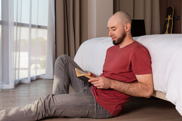 Portrait of a young, bearded, bald man is sitting on the floor at home reading a book. The concept of education and reading