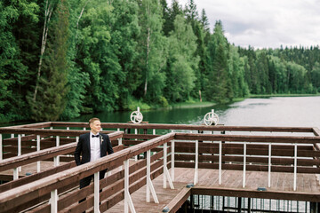 Groom keeps his hand in his pocket. Classic black tuxedo and details of the man's look at the wedding ceremony. The husband poses against the background of a forest lake.