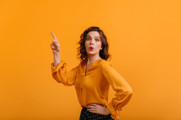 Shocked glamorous woman pointing with finger. Front view of amazed attractive girl isolated on yellow background.