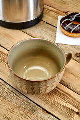 An empty brown mug stands on the table next to a doughnut and an electric kettle on a wooden background