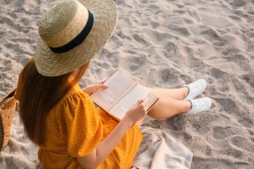 Young woman reading book on sandy beach, back view