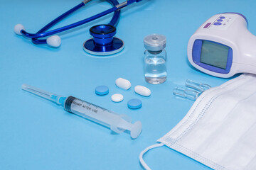 Fototapeta premium Ampoule, vial, syringe under white protective mask and non contact thermometer, stethoscope on blue background. Vaccination concept, disease prevention. Health care, Coronavirus vaccine development.