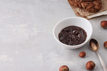 Chocolate nut spread in a white bowl with natural hazelnuts. Spoon, healthy cookies, hazelnuts on a light background. Vegetarian traditional breakfast close up. Copy space.
